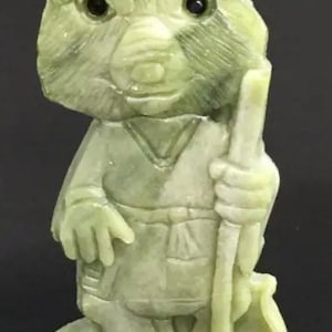 May include: A green jade figurine of a wolf wearing a robe and holding a staff. The wolf has black eyes and a detailed face.