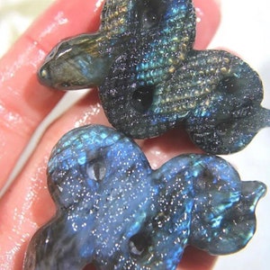 May include: Two iridescent labradorite gemstone snake sculptures. The snakes are carved in a detailed, realistic style and have a blue-green and black color.