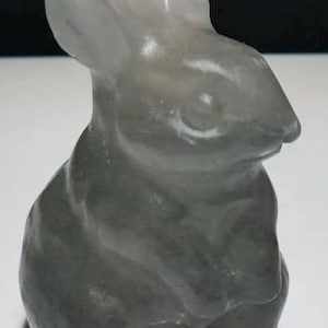 May include: A white glass sculpture of a rabbit sitting with its paws tucked under its chin. The sculpture is made of translucent glass and has a smooth, polished finish.