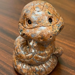 May include: A small, brown and gray stone sculpture of a snake with its head raised and eyes looking forward. The snake is coiled in a resting position.
