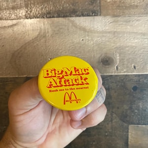 May include: A yellow button featuring the words "Big Mac Attack" in red, and the McDonald's logo. The button also includes the phrase "Rush me to the nearest".