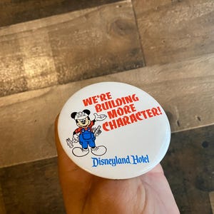 May include: White button with a cartoon image of Mickey Mouse wearing a hard hat and holding tools. The text on the button reads "We're building more character! Disneyland Hotel."