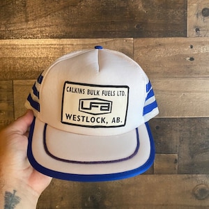 Peut inclure: Casquette de camionneur blanche avec des rayures bleues et blanches. La casquette a un patch qui dit "Calkins Bulk Fuels Ltd.  LFA Westlock, AB."