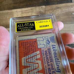 May include: A wrestling trading card encased in a clear plastic holder. The card features a wrestler illustration and text, including "KIMCHEE and K. UGANDAN HEAD". The holder has a yellow label with "ALL-STAR GRADING CO." and "CARD I.D. # 223281".