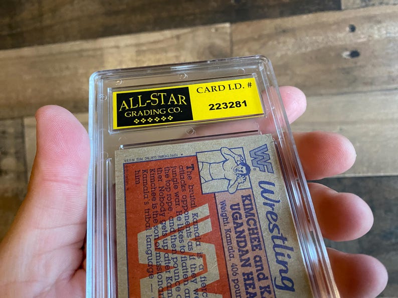 May include: A wrestling trading card encased in a clear plastic holder. The card features a wrestler illustration and text, including "KIMCHEE and K. UGANDAN HEAD". The holder has a yellow label with "ALL-STAR GRADING CO." and "CARD I.D. # 223281".