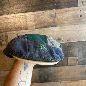Puede incluir: Una gorra plana de estilo patchwork con varios paneles de tela de mezcla de lana en tonos gris oscuro, carbón y verde bosque. La gorra incluye un pequeño detalle de trébol verde bordado en el lateral y un borde de color canela claro.
