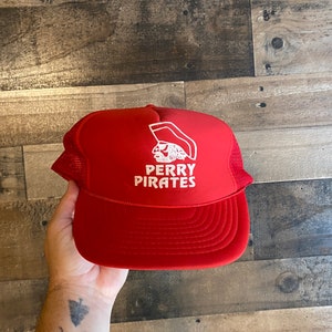 May include: Red mesh trucker hat with a white logo that says "Perry Pirates" and a cartoon pirate head wearing a hat.