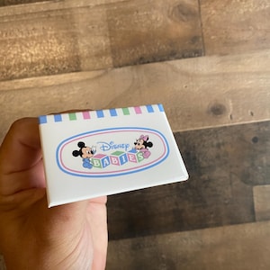 May include: A white box with a blue and pink striped border. The box has a logo that says "Disney Babies" with Mickey Mouse and Minnie Mouse in the center.