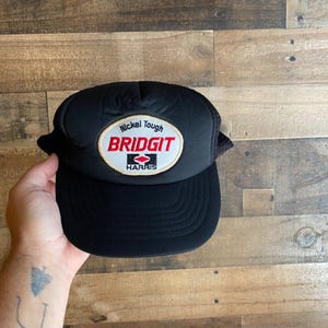 Puede incluir: Gorra de camionero negra de malla con un parche bordado en rojo, blanco y dorado que dice "Nickel Tough Bridgit Harris".