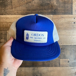 Può includere: Cappellino da camionista blu e bianco con una toppa bianca che dice "Ordos Fire Equipment Ltd. Edmonton"
