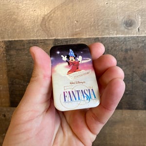 May include: A white and blue pin featuring Mickey Mouse as the Sorcerer's Apprentice from the movie Fantasia. The pin has a white background with blue stars and the text "Walt Disney's 50th FANTASIA" in a gold font.