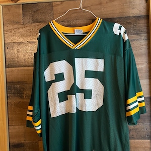 Pode incluir: Camisola de futebol verde com o número 25 em branco na frente. A camisola tem riscas amarelas e brancas nas mangas e na gola. A camisola está pendurada num cabide de arame.