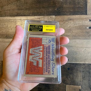 May include: A vintage wrestling card encased in a clear plastic holder. The card features the text "Wrestling Stars" and the names "Kimchee and Kamala the Ugandan Headhunter." The card is graded by All-Star Grading Co.