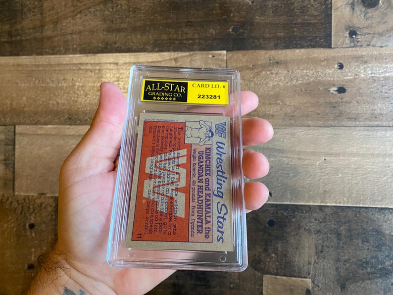 May include: A vintage wrestling card encased in a clear plastic holder. The card features the text "Wrestling Stars" and the names "Kimchee and Kamala the Ugandan Headhunter." The card is graded by All-Star Grading Co.