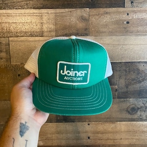 Peut inclure: Casquette de camionneur verte et blanche avec un patch blanc sur lequel est inscrit "Joiner Auctions" en lettres noires.