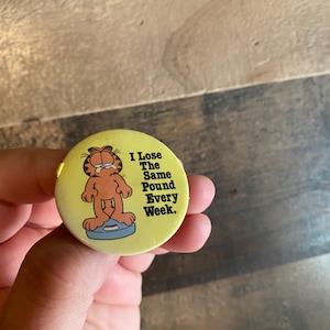 Op de afbeelding: Een gele ronde knoop met Garfield de kat op een weegschaal en de tekst "I Lose The Same Pound Every Week". De afbeelding is een humoristische kijk op gewichtsverlies. De knoop wordt in een hand gehouden.