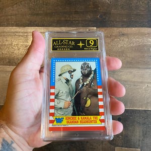 May include: A graded wrestling trading card featuring Kimchee and Kamala the Ugandan Headhunter. The card is encased in a clear plastic holder and has a blue, red, and white border. The card is graded NM or Higher.
