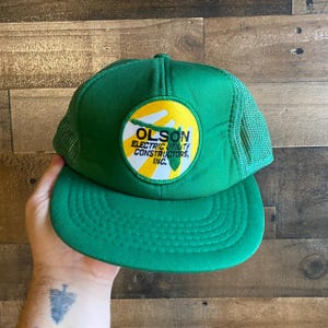 Peut inclure: Casquette de camionneur en maille verte et jaune avec un patch qui indique "Olson Electric Utility Constructors, Inc."