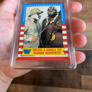 May include: A wrestling trading card featuring Kimchee and Kamala The Ugandan Headhunter. The card has a blue border with white stars and red and white stripes. The wrestlers are in costume, with one wearing a mask and the other with face paint.