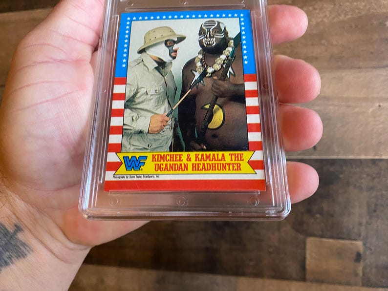 May include: A wrestling trading card featuring Kimchee and Kamala The Ugandan Headhunter. The card has a blue border with white stars and red and white stripes. The wrestlers are in costume, with one wearing a mask and the other with face paint.