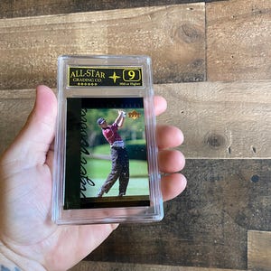 Puede incluir: Una tarjeta coleccionable calificada, encerrada en un soporte de plástico transparente. La tarjeta presenta a un golfista en acción, con el texto "Tiger Woods" y "All-Star Grading Co." visibles. La tarjeta está calificada con un 9.
