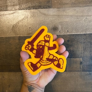 May include: Embroidered patch featuring a cartoon knight in maroon and gold, holding a sword. The patch has a yellow border and is designed with a playful, action-oriented pose. Ideal for jackets, bags, or other fabric items.