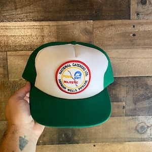 Peut inclure: Un chapeau de camionneur blanc et vert avec un patch circulaire qui indique "National Caterers Ltd. Norman Wells Pipeline" avec une carte stylisée d'un pipeline et les mots "Pedern, Majestic, NWTF" à l'intérieur de la carte.