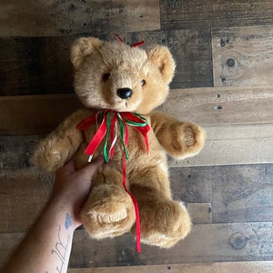 Puede incluir: Un oso de peluche marrón con una cinta roja, verde y blanca alrededor del cuello. El oso está sentado sobre una superficie de madera.