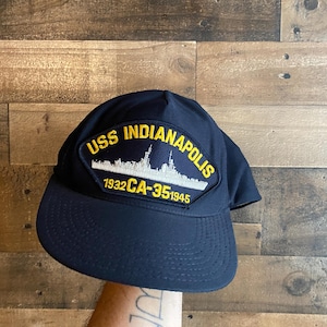 May include: A navy blue baseball cap with a patch displaying the text "USS Indianapolis" in yellow and white. The patch features a ship silhouette and the text "1932 CA-35 1945". The cap has a curved peak.