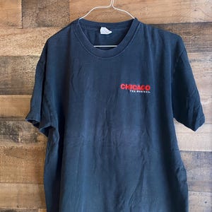 Pode incluir: Uma camiseta preta com as palavras "CHICAGO THE MUSICAL" em letras vermelhas.