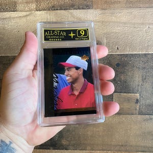 Puede incluir: Una tarjeta coleccionable calificada, dentro de un estuche de plástico transparente, que presenta a un golfista sonriente con un polo rojo y una gorra blanca. La tarjeta está calificada con un "9" por "ALL-STAR GRADING CO.".