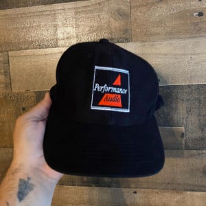 Puede incluir: Gorra de béisbol negra con las palabras "Performance Audio" en texto blanco dentro de un cuadrado blanco y rojo. La gorra está hecha de un material suave y tiene una visera curva.