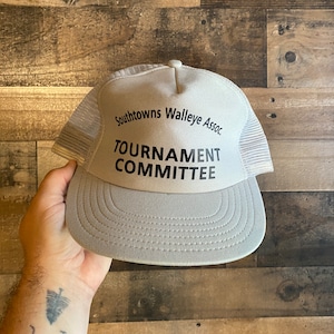 Puede incluir: Una gorra de camionero de malla gris y blanca con el texto "Southtowns Walleye Assoc. Tournament Committee" impreso en negro.