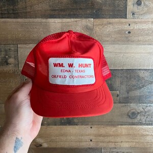 Puede incluir: Gorra de camionero roja de malla con un parche blanco que dice "WM. W. HUNT EDNA - TEXAS OILFIELD CONTRACTORS"