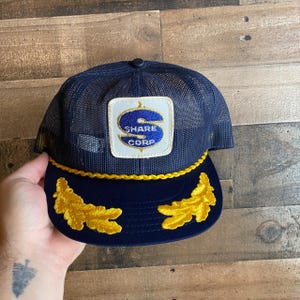 May include: A blue and yellow mesh trucker hat with a gold embroidered patch that says "Share Corp" and a gold embroidered design on the brim.