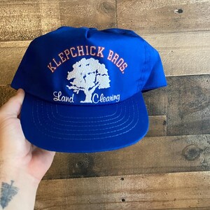 May include: Blue and red baseball cap with the text "Klepchick Bros. Land Clearing" and a white tree graphic.