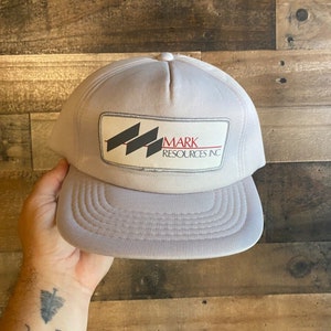 Peut inclure: Un chapeau de camionneur gris et blanc avec un logo noir et rouge qui dit "Mark Resources Inc."