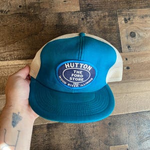 Op de afbeelding: Een blauwe en witte trucker-pet met een patch waarop staat "Hutton The Ford Store Inc. Wood River, Illinois".