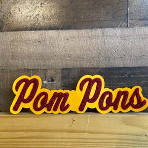 May include: A yellow felt patch with the words "Pom Poms" in maroon felt lettering.