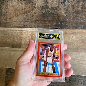 May include: A Michael Jordan basketball trading card, graded and encased in a clear plastic holder. The card features a photo of Jordan in his Bulls uniform, with the text "MICHAEL JORDAN" at the bottom. The card is graded "9" by All-Star Grading Co.