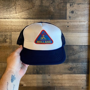 Peut inclure: Un chapeau de camionneur blanc et bleu avec un patch triangulaire qui dit "HOLLAND" et représente un moulin à vent.