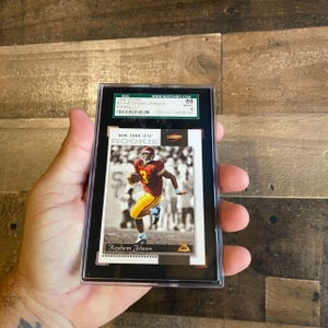 May include: A New York Jets rookie football card of Keyshawn Johnson, graded and encased in a black holder. The card features a photo of Johnson in his Jets uniform. The card is labeled "ROOKIE" and includes the text "NEW YORK JETS".