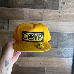 Può includere: Un cappello da baseball giallo con una toppa nera che dice "COYP LTD. CANADIAN OUTDOOR YELLOW PAGES".