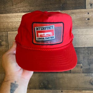 Peut inclure: Casquette de baseball rouge avec un écusson rectangulaire portant l'inscription "INTERSTATE CONSOLIDATION" et un dessin de deux semi-remorques. La casquette a une visière incurvée et un détail de corde tressée.