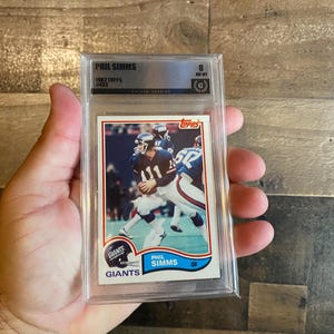 May include: A 1982 Topps Phil Simms football card, number 433, encased in a clear plastic protective slab. The card features a photograph of the player in a blue and white uniform, with the text Phil Simms, Giants, and the Topps logo visible.