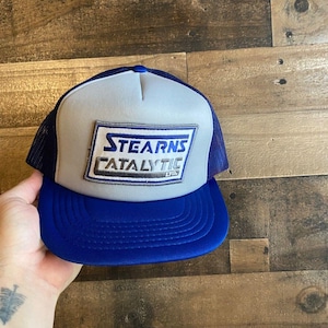 Peut inclure: Un chapeau de camionneur gris et bleu avec un patch blanc qui dit "Stearns Catalytic Ltd."