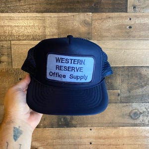 Peut inclure: Un chapeau de camionneur bleu marine avec un patch blanc qui indique "WESTERN RESERVE Office Supply".