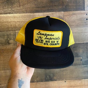 Peut inclure: Casquette de camionneur noire et jaune avec un patch qui indique "Lampman Imperials 80-81 Big Six 'A' Div. Champs".