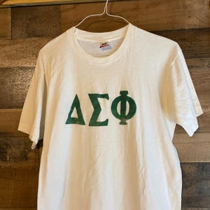 Puede incluir: Camiseta blanca con un logotipo verde de la fraternidad Delta Sigma Phi.