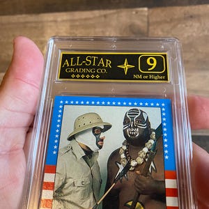 May include: A graded trading card featuring two wrestlers in masks. The card is encased in a clear plastic holder with a gold and black label that reads "ALL-STAR GRADING CO." and a "9" grade. The card has a blue border with white stars.